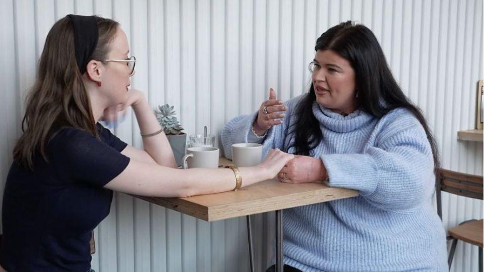 Kitty and Jessie sat at a table in a coffee shop. Kitty has long dark hair and is wearing a black headband. She has glasses and braces and is wearing a navy t-shirt. Jessie has long black hair and gold hoop earrings, and is wearing a light blue knitted roll neck jumper. 