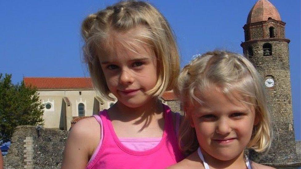 Laura and Gracie Nuttall stood together as young girls with a church behind them. Laura is on the left and has short, blonde hair and is wearing a pink vest. Gracie is to her right and a bit further forward and has the same short, blonde hairstyle