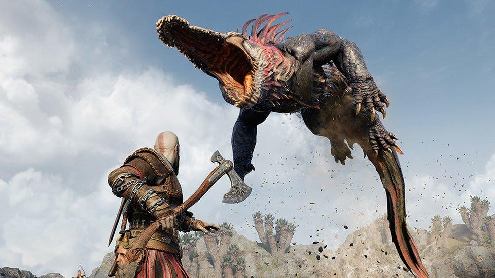 A screenshot from God of War Ragnarök shows main character Kratos, viewed from behind, in a battle stance. He's readying his axe as a giant, crocodile-like creature leaps into the air, its jaws wide open, exposing rows of razor-sharp teeth as it prepares to attack our hero. Its long body is arched, and its long tail brushes against the ground, where a cloud of dust and debris has been kicked up as it left the ground moments before.