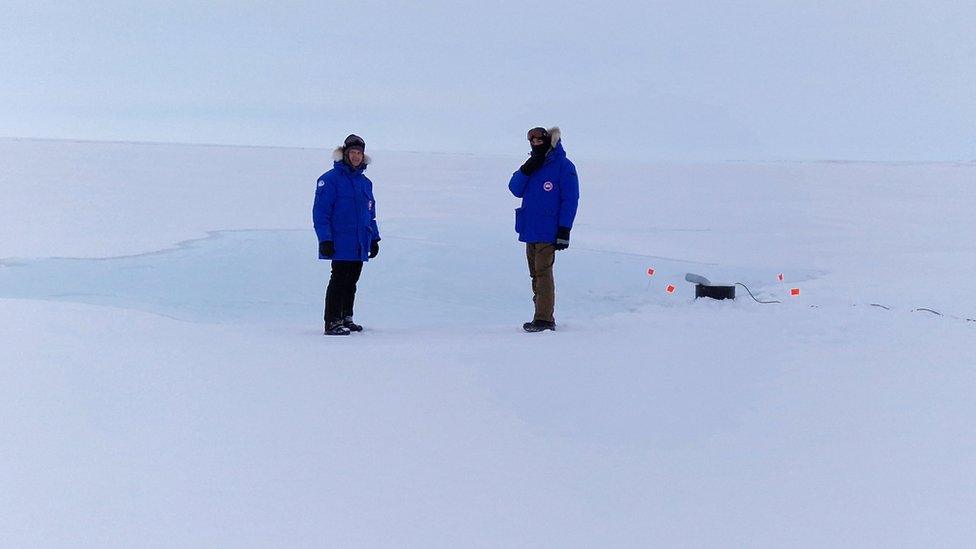 Two researchers are wrapped up in Arctic gear as they watch the seawater pump in action