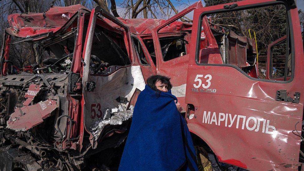 A woman standing in front of a wreckage of a fire engine with a blue blanket partially covering her face