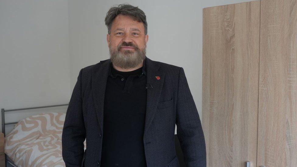 Richard Priest with a beard wearing a black blazer and black T-shirt standing in front a bed and a wardrobe in the social housing