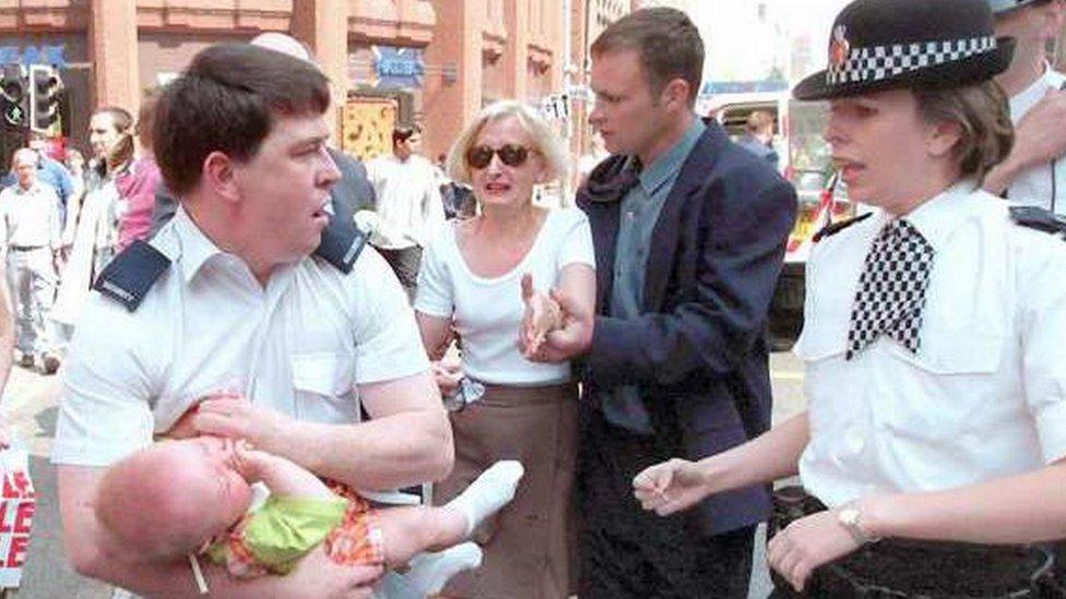 Security guard clutches injured baby Sam Hughes with family and policewoman looking on