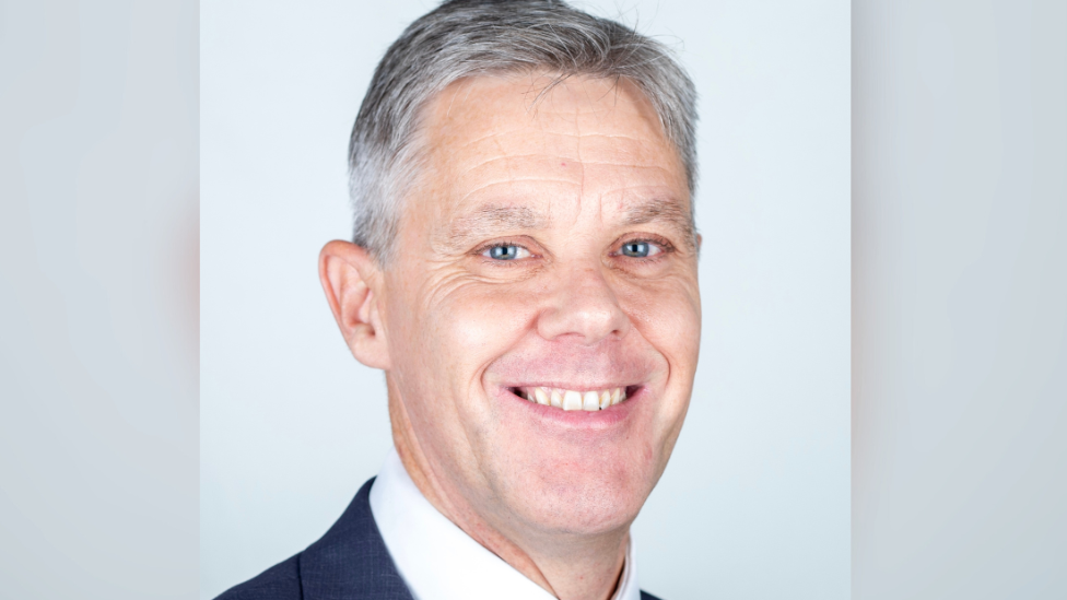 Matthew Warren wearing a dark suit, white shirt and tie looking towards the camera. He has grey hair and bright blue eyes. He is standing against a plain light blue background.