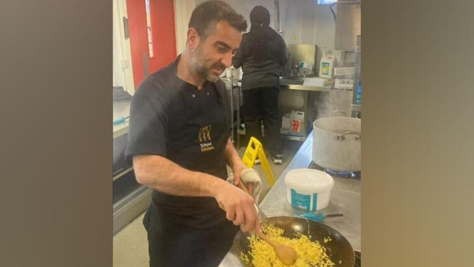 Man with a wok of rice cooking in a school kitchen. Another woman is behind preparing food with her back to the camera. Both people re wearing black t-shirts, trousers and aprons.