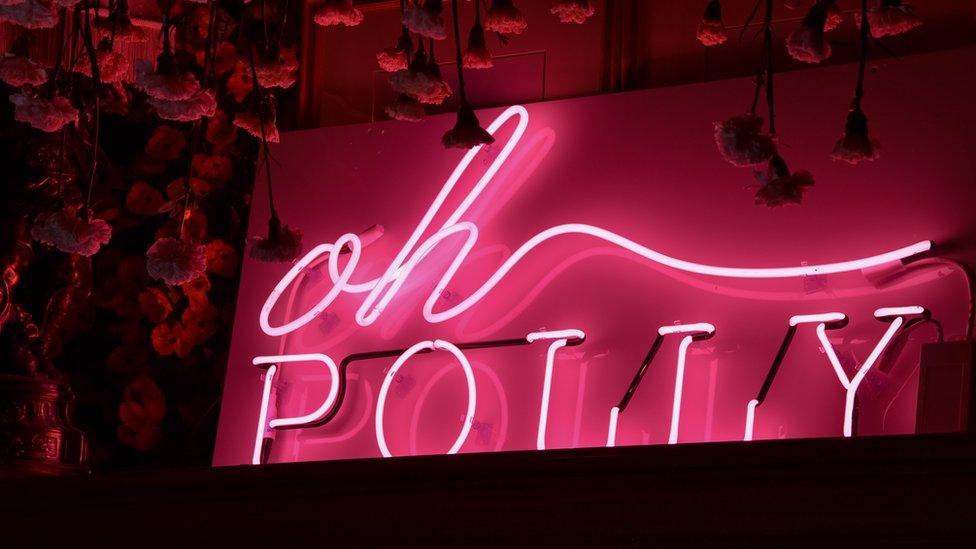 A pink fluorescent sign of the Oh Polly logo against a black backdrop.