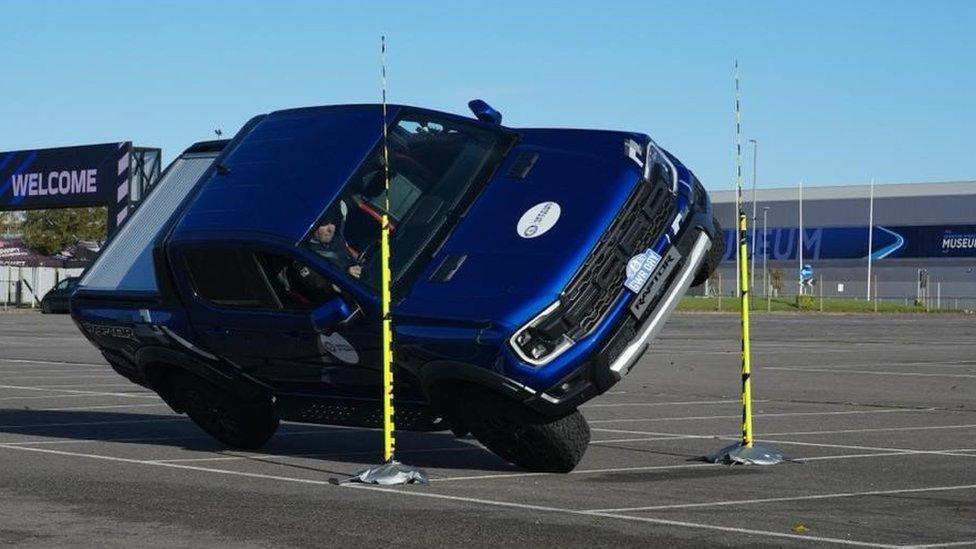 A blue pickup tilted on two wheels drives between two yellow poles