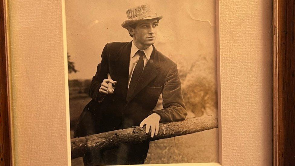 Old photo of Tony James. It is black and white. He is leaning over a fence, wearing a suit and a hat.