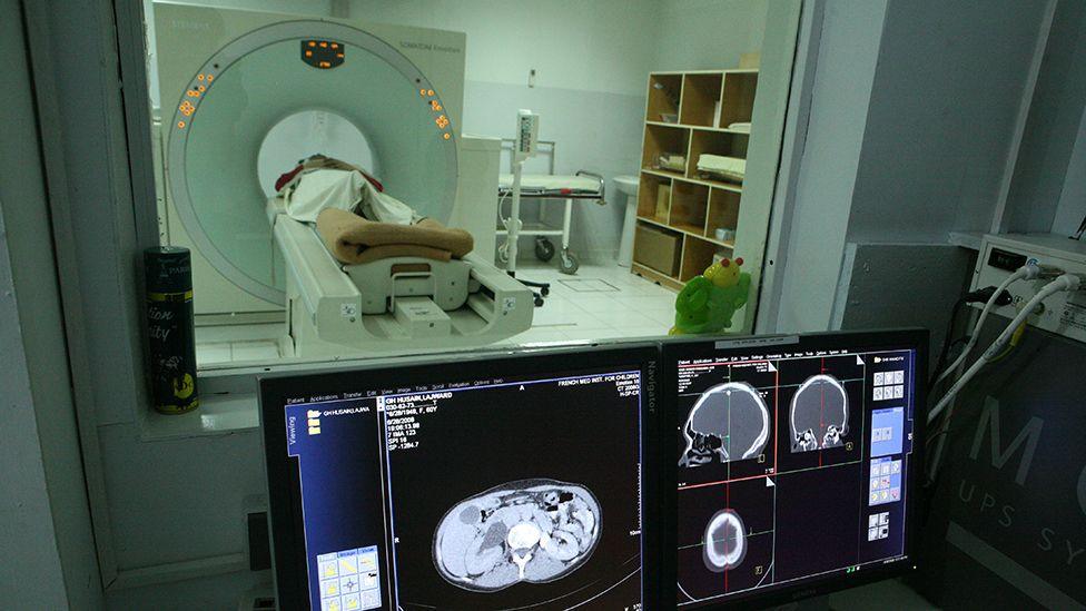 A person having their head scanned in a CT scanner in the background with X-ray images of their brain displayed on computer monitors in the foreground