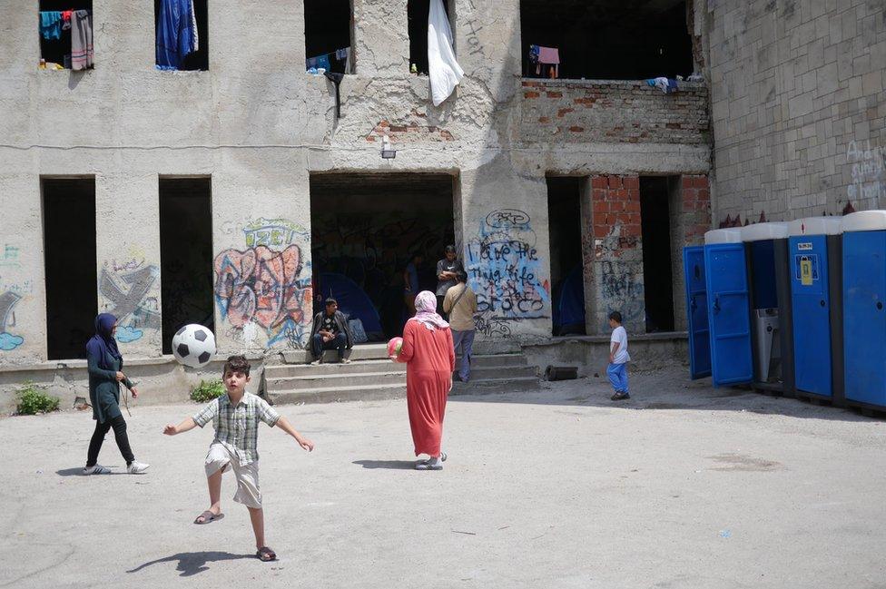 A young boy plays with a ball in front of the building, which has no doors or windows - a few portable toilets stand against a nearby wall