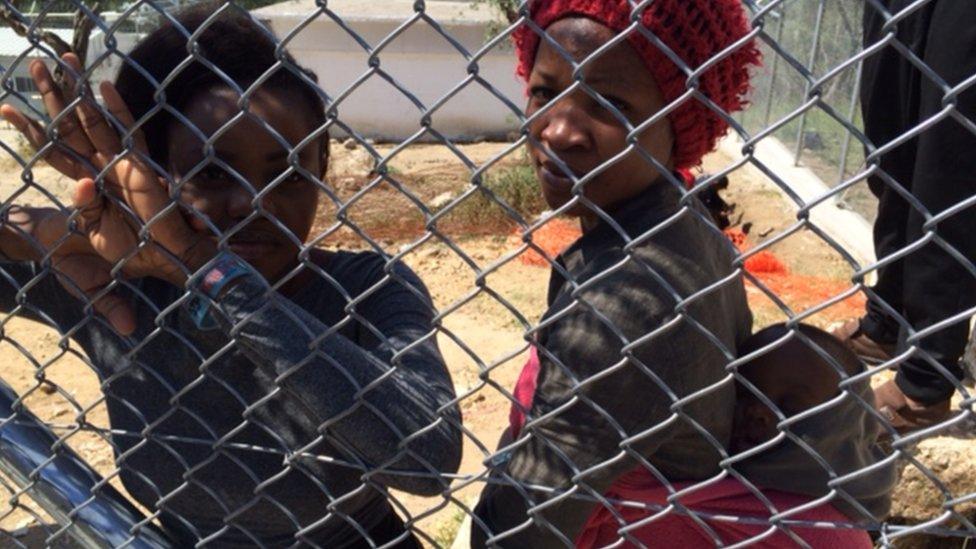 Blessing, a migrant woman, stands behind a fence with her infant son