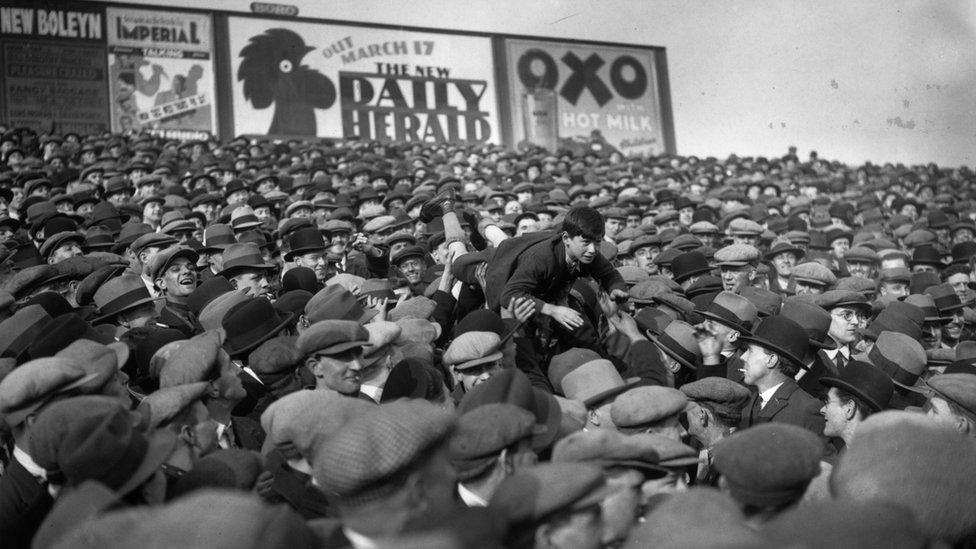 Fans pass a young boy over their heads to the front so that he can see the game better, 1 March 1930