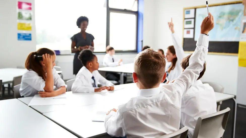 Generic view of school children in white shirts. A child with his back to the camera has his hand in the air.