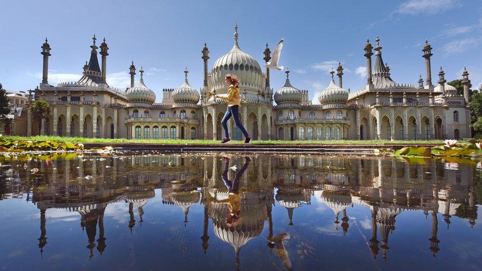 A teenage girl reflected from across a pond - runs pursued by a seagull past the seaside town's landmark Georgian Royal Pavilion with it's famous domes and minarets