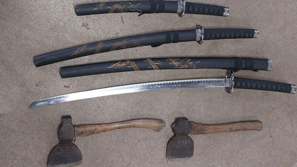 Six weapons lying on the ground. Three swords have black sheathes with gold decorative work, the fourth is an unsheathed sword and beneath them are two axes.