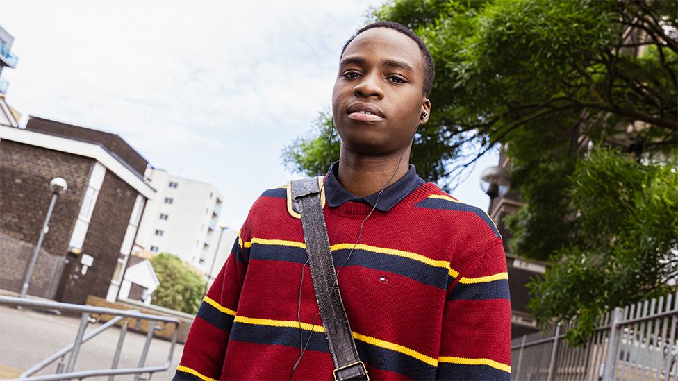 Character of Bayo wearing a maroon sweater with blue and yellow lines. He is looking at the cmaera and has a bag strap going across his top. The background has buildings, silver railings and a large green tree in the top right.