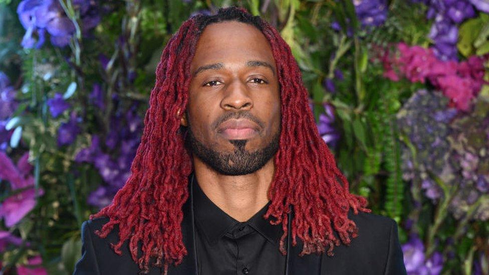 Avelino is pictured at Stormzy's 30th Birthday at The Biltmore Mayfair on July 28, 2023 in London. Avelino is a black man in his 30s and he has his hair in red braids just past his shoulders and a short styled beard. He is wearing a black suit and shirt and is looking straight at the camera - he is not smiling and has his mouth closed as he tilts his chin up. He is pictured in front of a backdrop of green foliage interspersed with pink, purple and blue flowers.