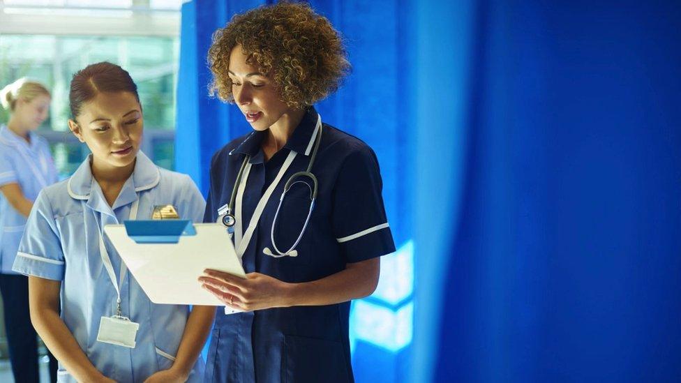 A stock image of two nurses standing on a hospital ward chatting. They are looking at a clipboard.