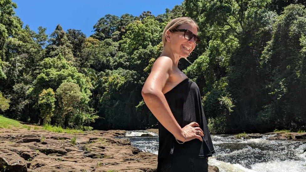 Emma Lovell in a black top and shorts standing at the base of a waterfall.