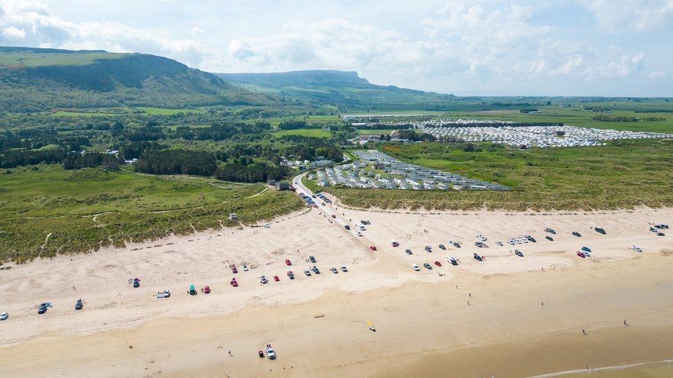 An aerial shot of Benone beach on a sunny day - cars are parked on the sand while behind the beach caravan parks can be seen