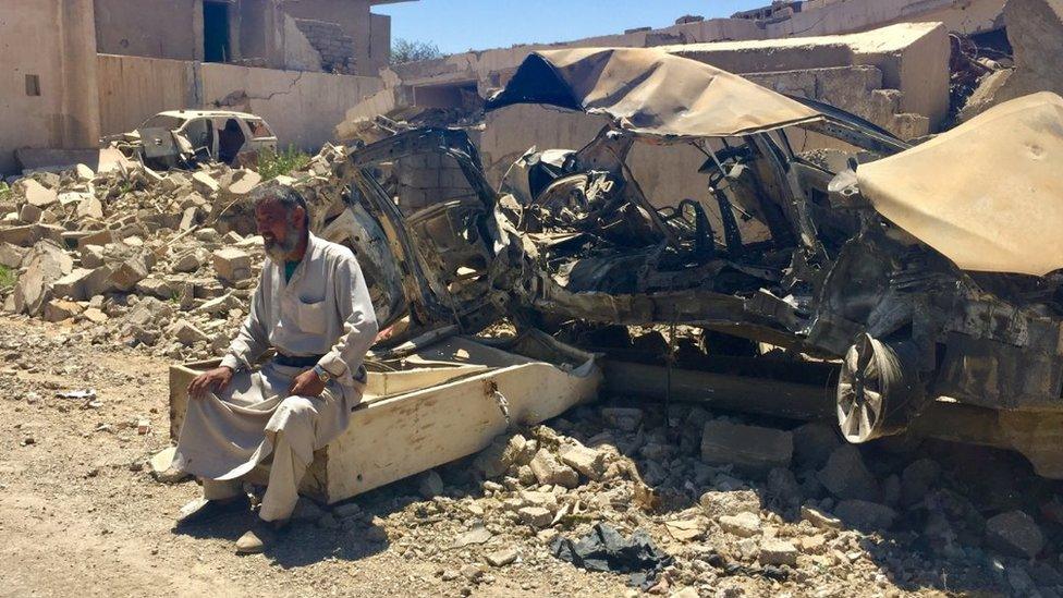 A man sits in front of a burnt out car