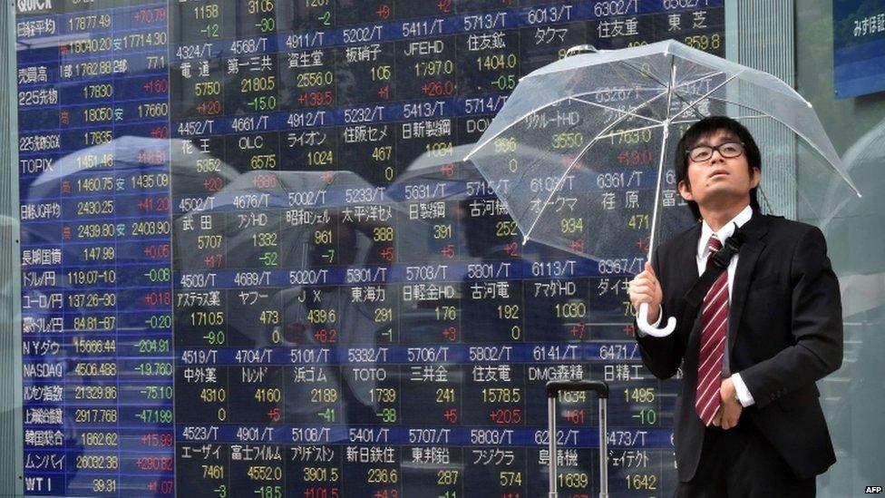 A businessman with an umbrella looks up to the rainy sky as he passes a share prices board in Tokyo on August 26, 2015.