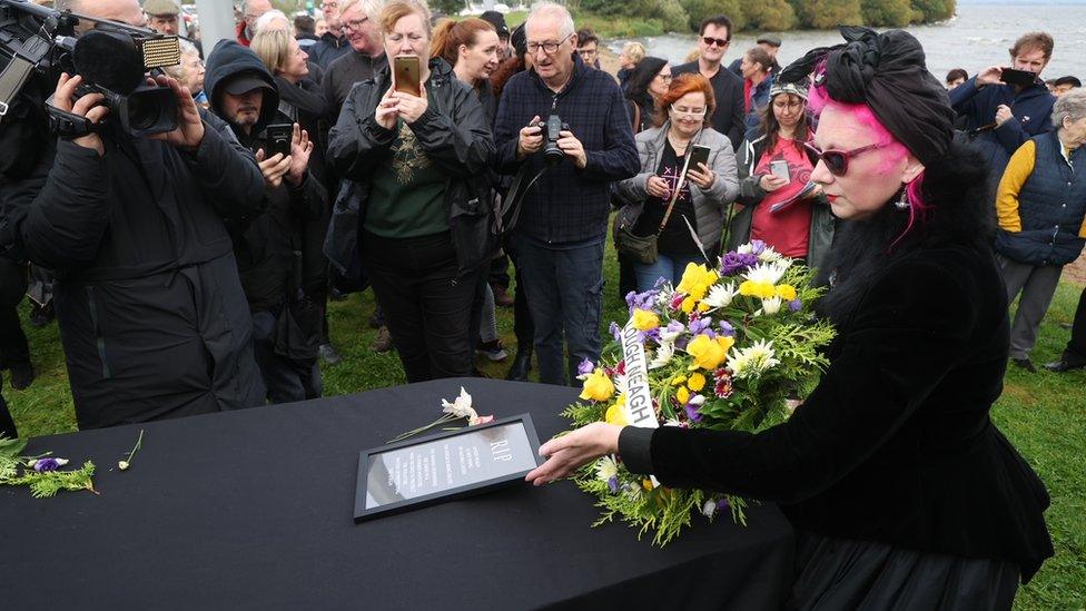 A woman dressed in black lays a wreath on a black-covered coffin