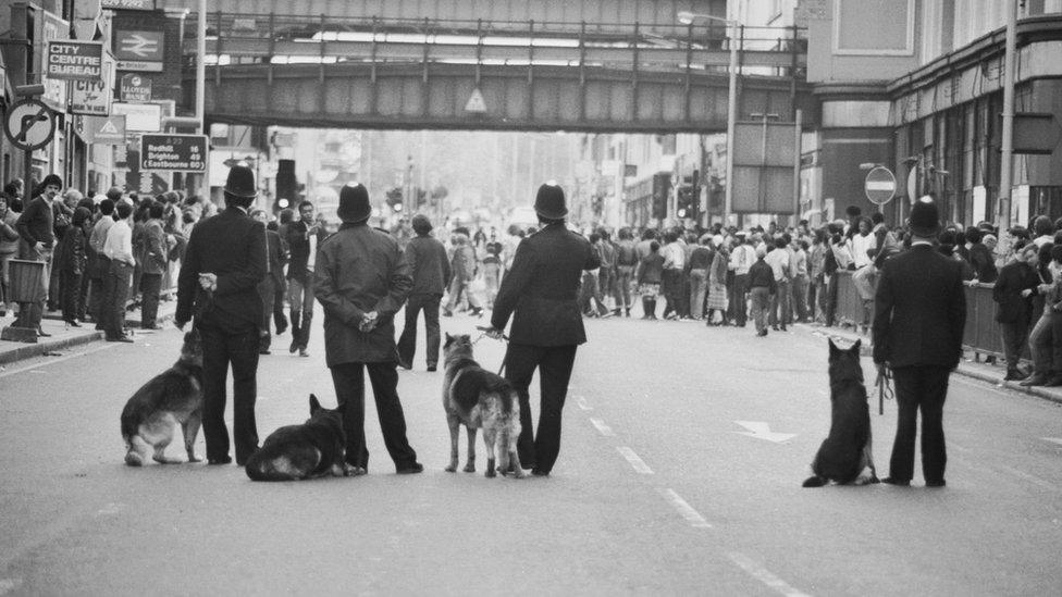 Four police officers stand facing a crowd of people a few hundred yards away