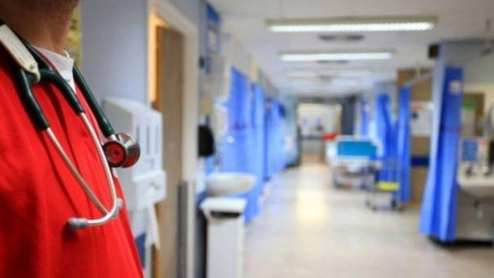 A doctor stands on a hospital ward. He is wearing a stethoscope around his neck and red theatre scrubs.