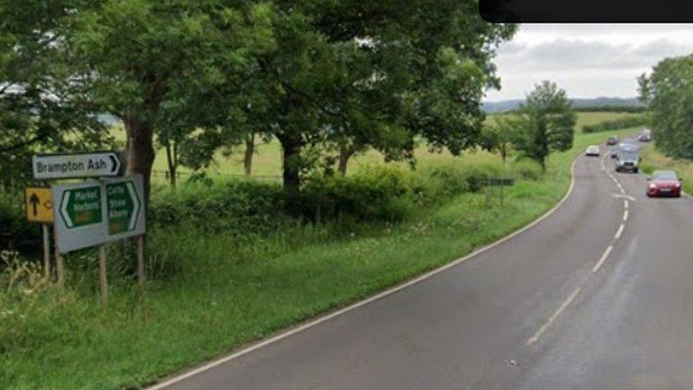 Main road through rural area with sign saying "Brampton Ash". Trees on both sides.