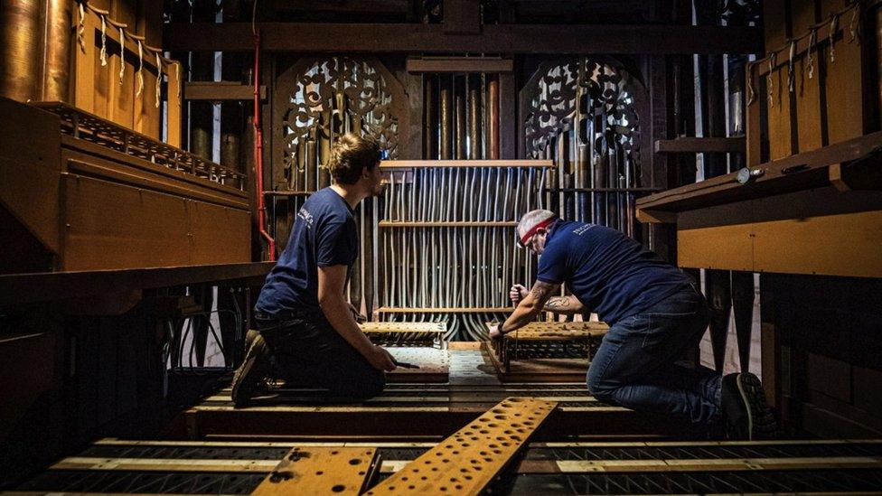 Two men refurbishing the Gloucester Cathedral organ. They are kneeling down and using tools to work. There are two planks of wood laid out on the floor next to them.