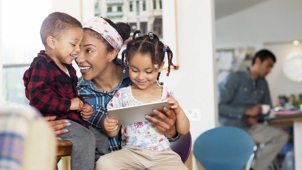 A woman plays with her two children at home with her partner in the background