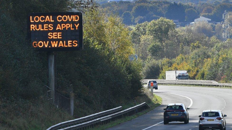 A photograph of a sign that says "Local Covid rules apply, see gov.wales", taken along a road during the Covid pandemic