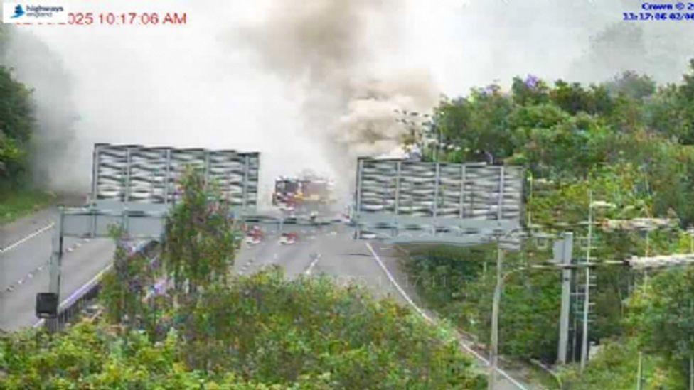 A blurry photo from a traffic camera showing billowing smoke coming from the side of a motorway, behind gantry signs. Lots of foliage in the foreground. A fire engine is at the scene.