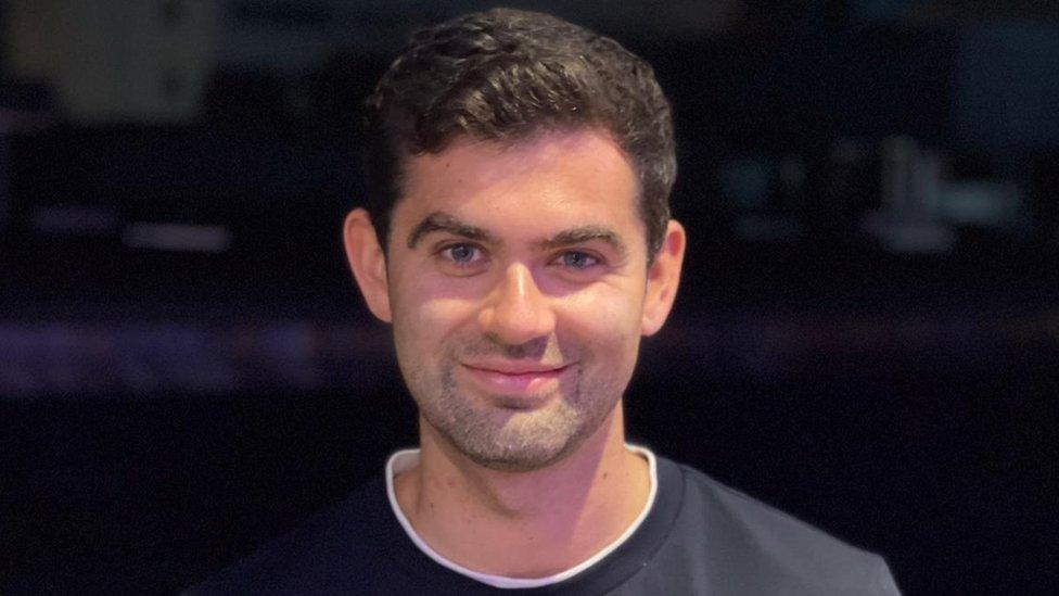 Al Smith smiles, wearing a navy t-shirt. He's against a dark background in what looks like a studio setting. He's got one eyebrow slightly arched, and sports neat, slightly curly black hair and light