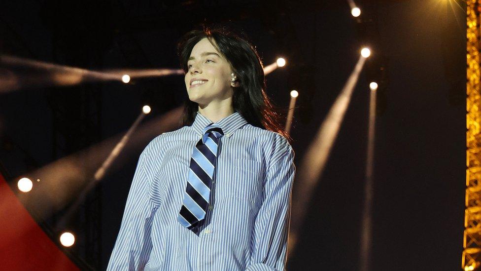 Billie Eilish performs onstage during Global Citizen's Power Our Planet: Live in Paris on June 22, 2023 in Paris. Billie is a 22-year-old white woman with long black hair and green eyes. She wears a blue and white striped shirt with a blue and navy striped tie. She smiles, looking to the right of the camera. Behind her, spotlights can be seen against a dark sky and the Eiffel Tower, lit up, is just to the right of the frame