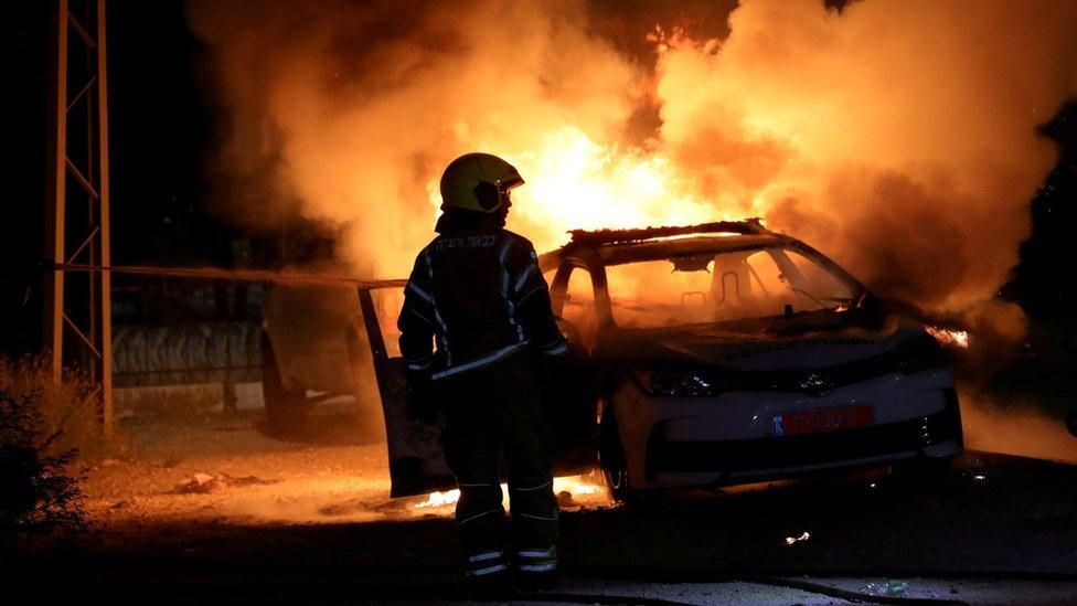 An Israeli firefighter stands near a burning Israeli police car during clashes between Israeli police and members of the country"s Arab minority in the town of Lod, Israel (12 May 2021)