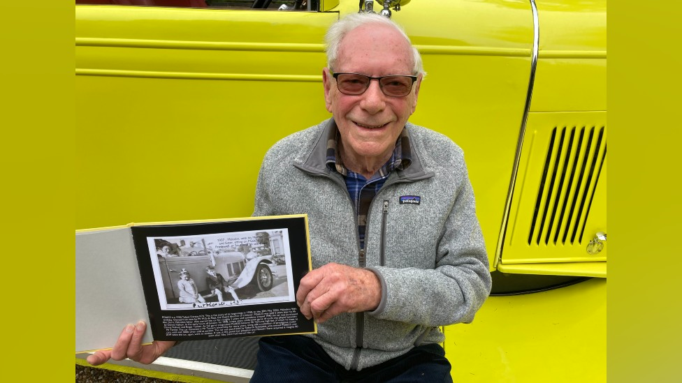 Malcolm Stern holding a family photograph whilst sitting on the running board of the car