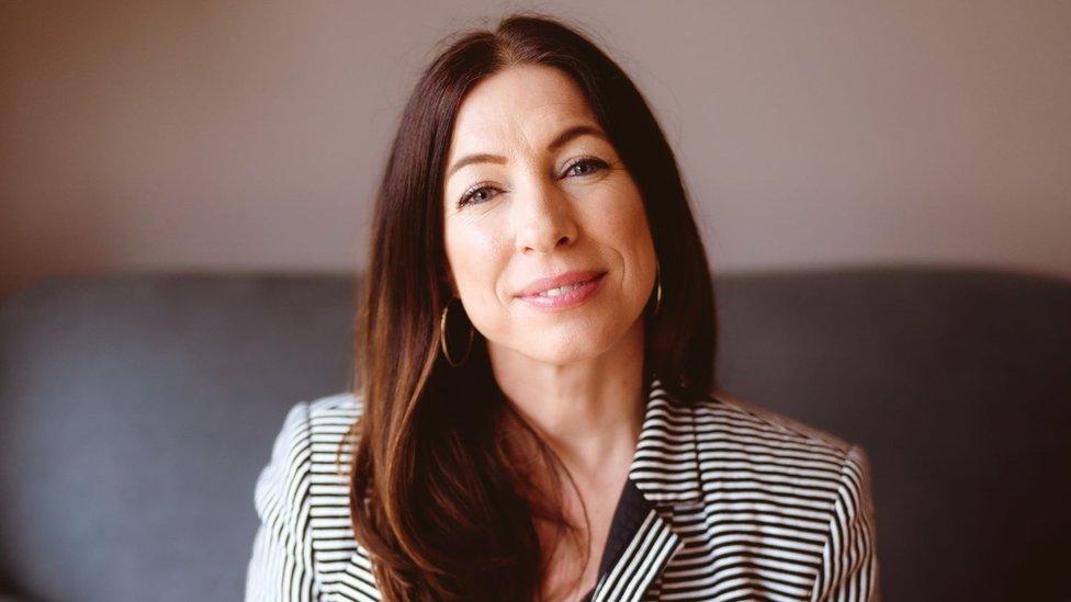 Dr Beth Mosley. Dr Beth is a white woman in her 40s with long brown hair which she wears loose over her right shoulder. She has blue eyes and is smiling at the camera with her head tilted slightly to her right. She wears a striped black and white blazer and hooped earrings. She is photographed inside on a grey sofa in front of a muted pink wall.