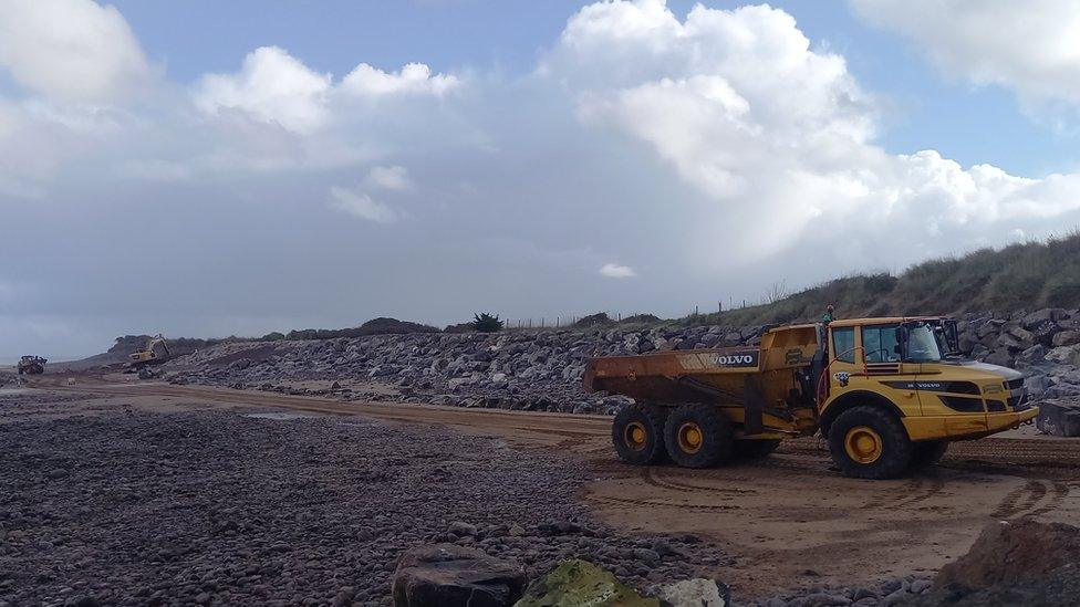 A yellow dumper truck delivering large granite boulders on a sandy beach on a sunny day