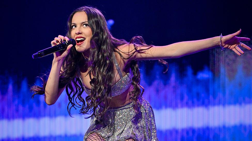 A young woman holds a microphone to her mouth and leans forward slightly, causing her long black hair to hang down in front of her. Her other arm is outstretched, as if she's trying to encourage the crowd to sing along with her. Behind her an elaborate arc of stage lights glows bright blue.