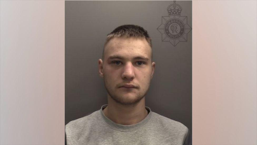A police head and shoulders shot of a man with short blond hair and shorter sides looking into the camera with a serious expression. He is wearing a grey jumper.