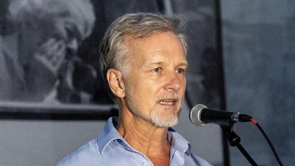 Paul Lowe stands in front of a microphone giving a speech at a photography exhibition 