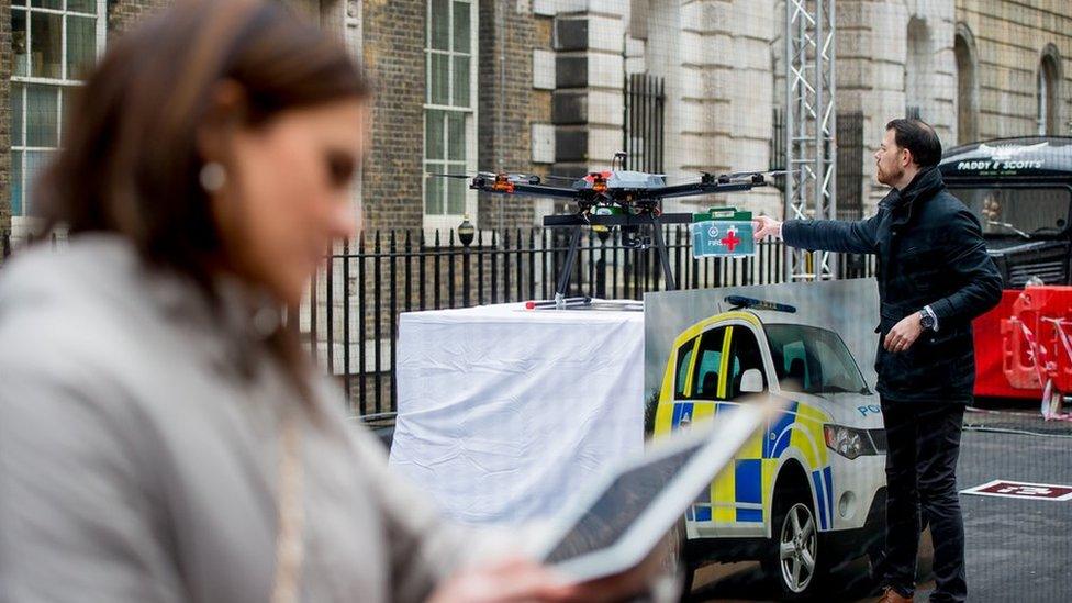 A woman watches on a tablet as a man loads first aid supplies onto a drone at a trial in central London