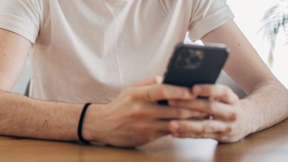 Generic image of a man in a white T-shirt holding a smartphone in front of him on a wooden table.