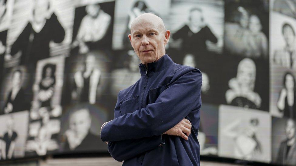 A portrait of a man with the backdrop of an exhibition behind him.