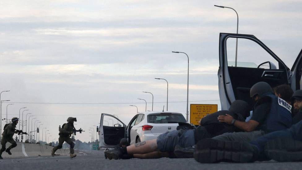 Journalists take cover behind cars as Israeli soldiers take position during clashes with Palestinian fighters near the Gevim Kibbutz, close to the border with Gaza on 7 October, 2023