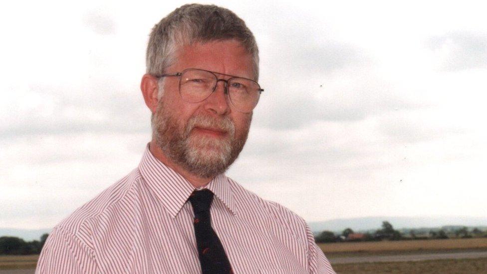 A man in a shirt and tie with a brown and grey beard and glasses