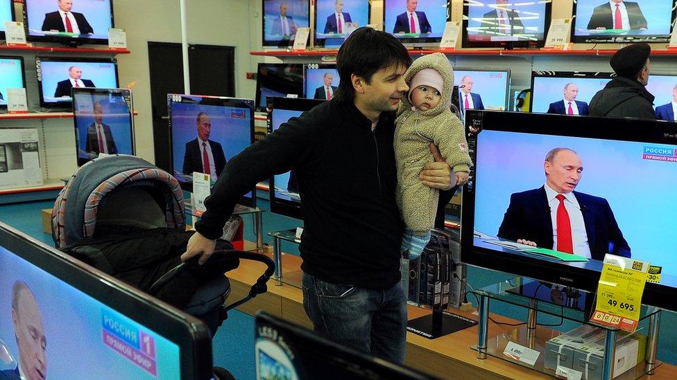 A man holds a baby as he walks past a bank of televisions showing Vladimir Putin