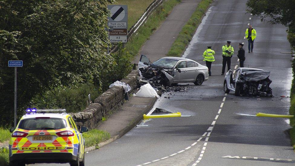 Two cars have crashed on a road. One is partially sitting on a footpath, the other is in the middle of the road. Both are damaged. Police are on the road.